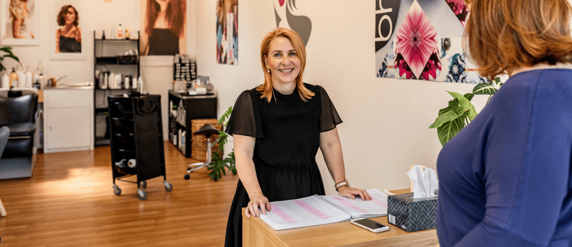 A lady with shoulder length straight red hair, wearing a black short sleeved dress is standing behind a check-in desk in a hairdressing salon. She is smiling and ready to help customers.